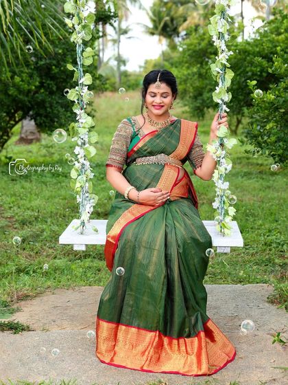 A joyful outdoor portrait of a mother-to-be in a green and red silk saree, seated on our floral swing with bubbles floating around.