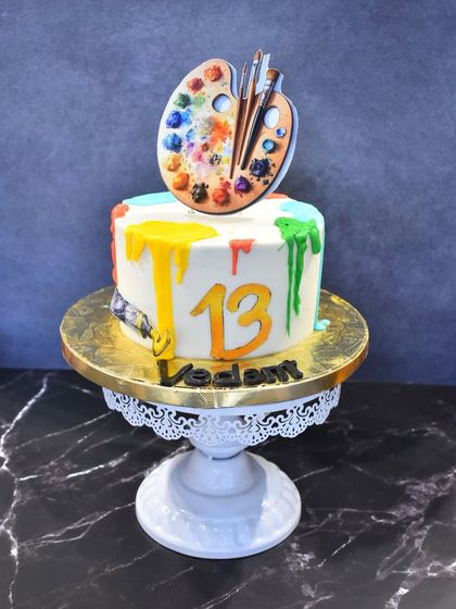 A colorful cake for a young artist turning 13. The cake is topped with an edible paint palette and brushes, with vibrant paint drips running down the sides.
