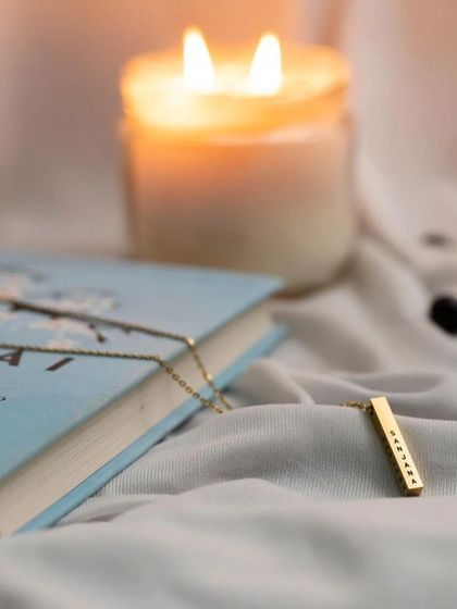 A gold bar pendant styled in a cozy setting with a book and a candle. This image associates the jewelry with moments of relaxation and personal reflection.