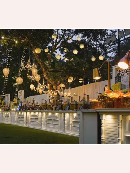 A wide shot of our white buffet counters illuminated at night, with chandeliers and lanterns hanging from the trees, blending functional food service with stunning decor.