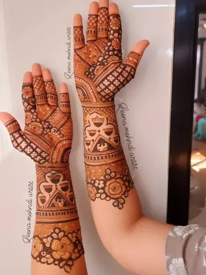 This design shows how different patterns on each hand can still create a cohesive bridal look. One arm features jhumka motifs while the other has a grid pattern, tied together by floral cuffs.