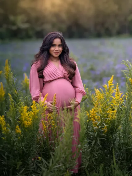 An expecting mother stands in a field of yellow and purple wildflowers, her pink dress complementing the natural colors. This outdoor portrait is vibrant, soft, and full of life.