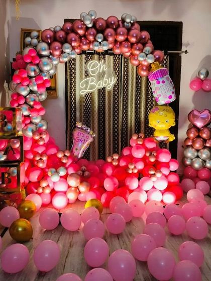 An 'Oh Baby' shower setup announcing "It's a Girl!". The room is filled with pink balloons, and the backdrop features a circular arch of pink and silver balloons with a neon sign.