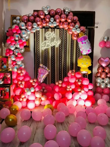 An 'Oh Baby' shower setup announcing "It's a Girl!". The room is filled with pink balloons, and the backdrop features a circular arch of pink and silver balloons with a neon sign.