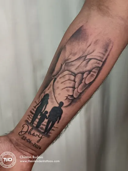 This forearm tattoo beautifully merges multiple symbols of a father's love. It combines a silhouette of his family, his son's name and birthdate, and the image of a father's hand holding his child's.