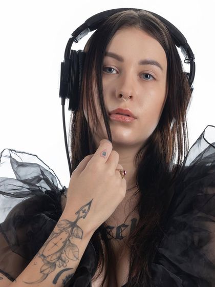 A moody portrait of a female DJ with headphones, tattoos, and a sheer black top, captured against a clean white background.