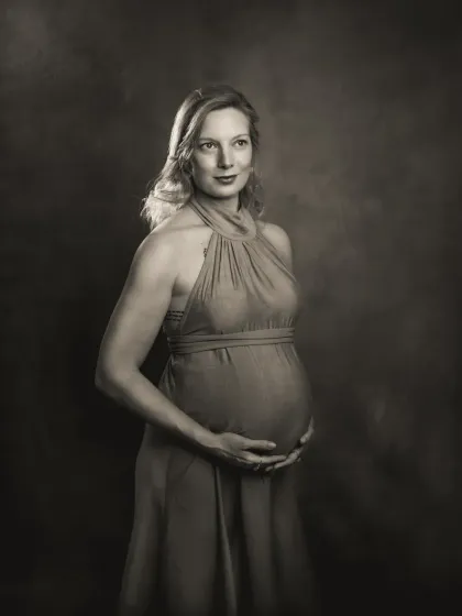 A duplicate of ID 19, this is a contemplative pose from a studio maternity session. The expecting mother looks down at her belly, her hands cradling it gently, against a painterly, textured backdrop. This is the B&W version.