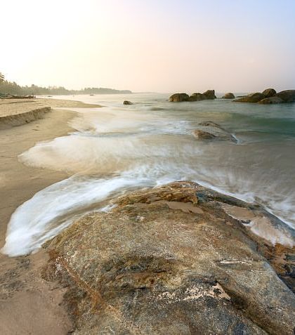 A long exposure captures the white foam of a wave receding from a sandy beach, with a large rock as the anchor point of the composition.