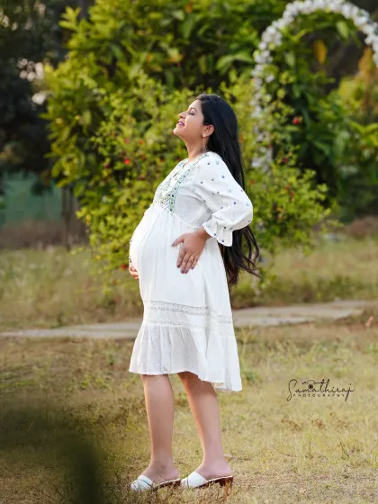 A serene outdoor portrait of a mother-to-be enjoying the sunlight. Her simple white dress and the lush green background create a fresh, natural, and peaceful image.
