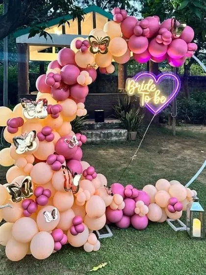 An outdoor 'Bride to be' garden party with a semi-circular balloon arch in shades of pink and peach, and a heart-shaped neon sign.