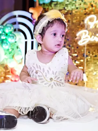 A baby girl sits in front of a glittering gold sequin backdrop with a "Happy Birthday" neon sign, a perfect setup for a glamorous first birthday shoot.