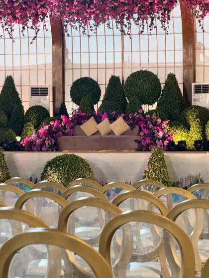 A gorgeous seating area set against a backdrop of a greenhouse-style window. The space is filled with sculpted topiaries and a profusion of purple and pink flowers.