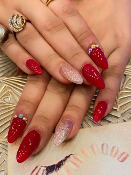 A stunning set of bright red, almond-shaped nails perfect for a party. Two nails are accented with a light pink glitter base, while others have small crystal clusters at the cuticle.
