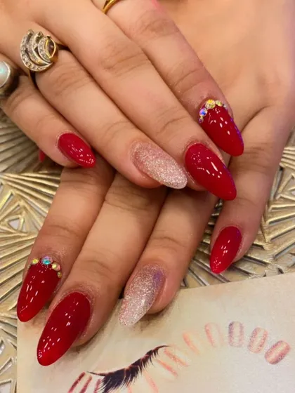 A stunning set of bright red, almond-shaped nails perfect for a party. Two nails are accented with a light pink glitter base, while others have small crystal clusters at the cuticle.