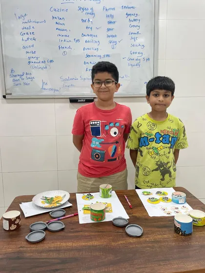Two students proudly stand by their finished cooking game. This activity combines play with learning, as they use vocabulary related to food and cooking while working together on a fun project.