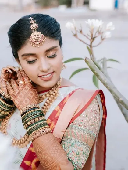 A serene bridal portrait, with the bride adjusting her earring. The natural outdoor setting and her beautiful white saree create a peaceful and elegant moment.