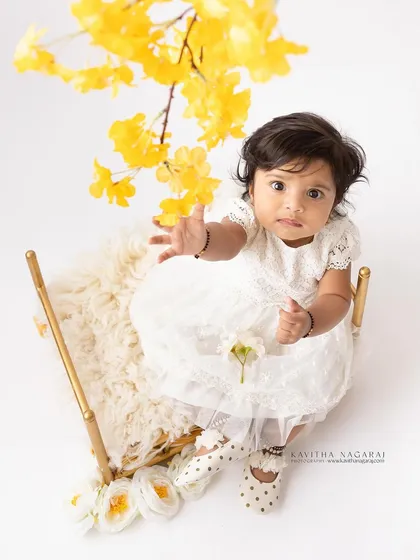 A six-month-old girl reaches for the camera from her seat. The high-angle shot and bright lighting create a fresh and engaging portrait.