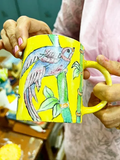 A hand-painted ceramic mug featuring a colorful bird on bamboo. We offer on-glaze ceramic painting workshops for creating functional art.