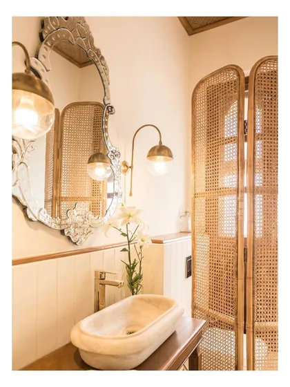 The powder room at Loto Bianco showcases a blend of rustic and refined elements. A carved marble sink sits atop a wooden vanity, complemented by a hand-carved mirror, brass light fixtures, and a woven cane screen for privacy.