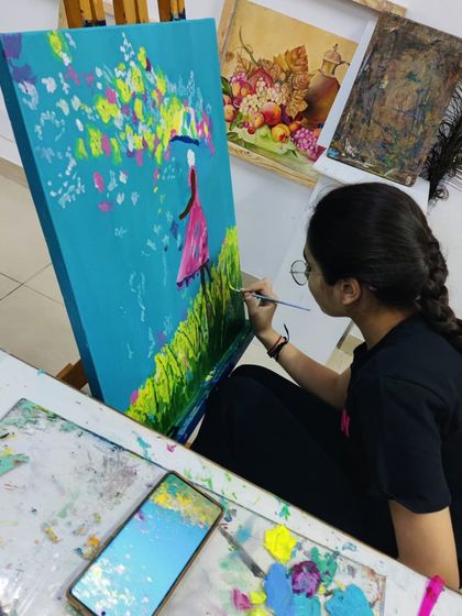 A student works on an imaginative painting of a girl with an umbrella in a field, using a palette knife and brush to create texture.