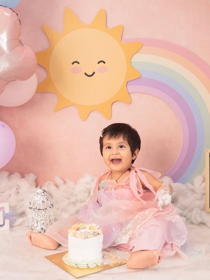 The pure delight of a first birthday cake smash. The colorful rainbow backdrop and fluffy clouds make this "You Are My Sunshine" theme a cheerful setting for a memorable milestone.