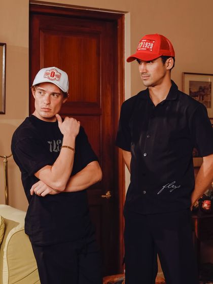 A candid-style shot of two models wearing branded caps and t-shirts. The relaxed, indoor setting gives the apparel a casual, lifestyle feel, making it look like part of an everyday wardrobe.