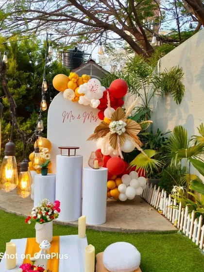 A bright and cheerful anniversary or birthday setup in my garden. This design features a custom white panel with "Mr & Mrs", a vibrant red and yellow balloon arrangement, and a low picnic table for guests.