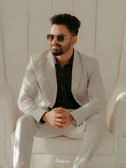 A dapper solo portrait of the groom-to-be, looking sharp in a light-colored suit while seated in a modern white throne chair.