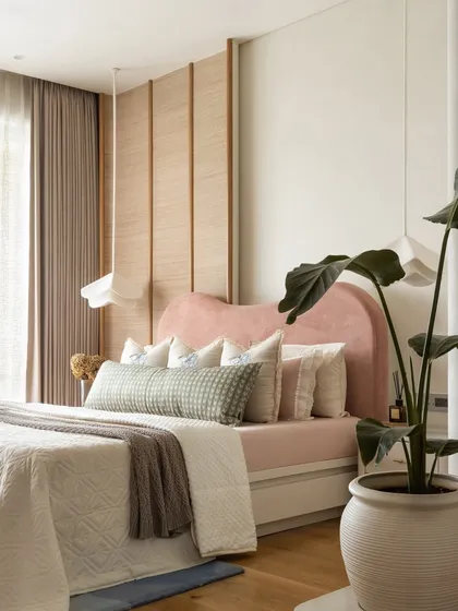 This master bedroom is a serene cocoon, anchored by a custom-designed curved headboard in a subtle peachy pink. A tactile banana fibre panel partially frames the wall, adding organic charm and texture, while bespoke pendant lights cast a delicate, intimate glow.