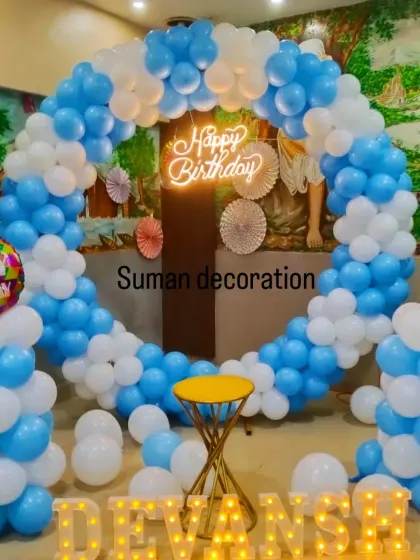 A simple and bright setup for a boy's first birthday. It features a blue and white balloon ring, a neon sign, and the name 'Devansh' in marquee letters.
