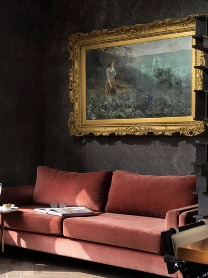 A cozy corner near the dining area, where an ash-pink sofa offers a comfortable spot for a pre-dinner drink. The space is defined by a dark, textured wall and a beautiful painting in a gilded frame, creating a moody and intimate atmosphere.
