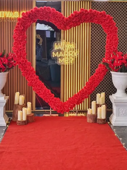 An elegant indoor proposal decoration featuring a bold red floral heart arch against a modern, textured wall. A red carpet runner and classic white urns filled with red flowers complete this sophisticated and passionate setup.