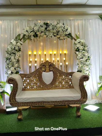 A simple yet beautiful half-saree program setup in JP Nagar. The stage uses a white floral arch, a classic golden sofa, and green turf flooring for a fresh look.