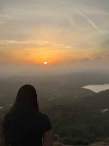 Another beautiful shot of a trekker watching the sunrise at Uttari Betta, with a lake visible in the distance.