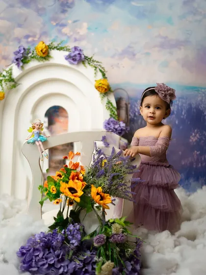 A sweet portrait of a baby girl in a purple dress, surrounded by flowers. The whimsical setup creates a magical, fairytale-like atmosphere.