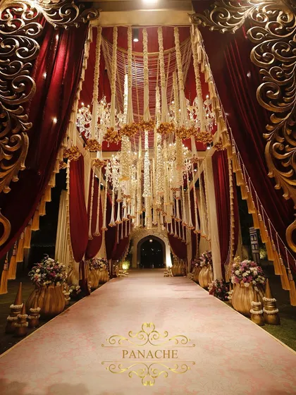 This grand walkway, draped in rich maroon velvet and framed by intricate gold panels, sets a regal tone for the wedding. The ceiling of hanging Rajnigandha tassels adds a traditional fragrance and texture.
