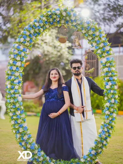A stylish couple poses within a large floral arch, blending modern and traditional elements in their attire.