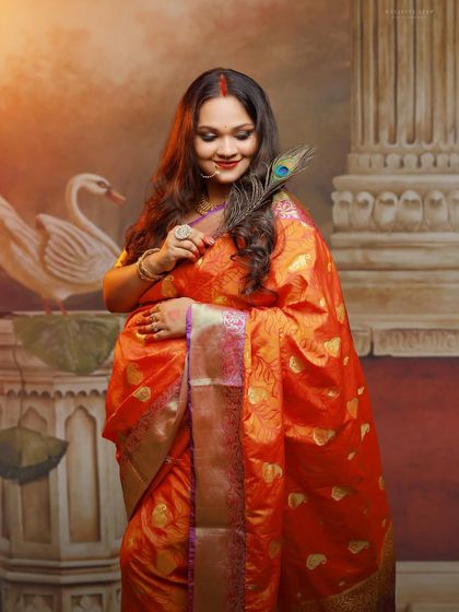 A graceful solo portrait of a mother-to-be in a vibrant orange Banarasi saree. The peacock feather adds a touch of tradition and symbolism to this elegant studio shot.