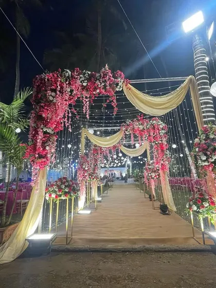 A beautiful floral entrance for an evening wedding. The combination of pink and red flowers, draped fabric, and warm fairy lights creates a romantic and inviting pathway for guests.