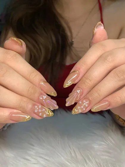 A beautiful set of stiletto nails with a nude base, gold chrome tips, and delicate 3D shell and pearl accents.