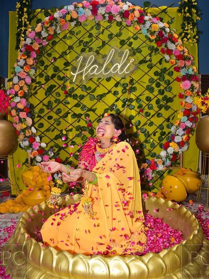 Capturing pure joy. A happy bride enjoying her floral shower in a custom Haldi setup I designed, featuring a 'Haldi' neon sign against a green trellis backdrop.