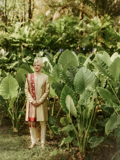 A portrait of the groom, William, standing amidst lush tropical foliage in Thailand, the vibrant green making his golden sherwani pop.