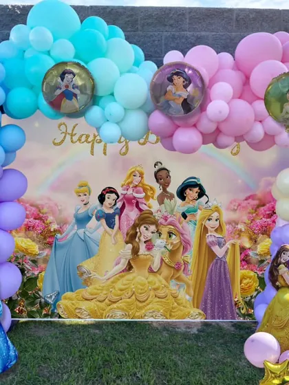 A full and vibrant Disney Princess backdrop for an outdoor party. The setup features a large character poster and a multi-colored balloon arch.