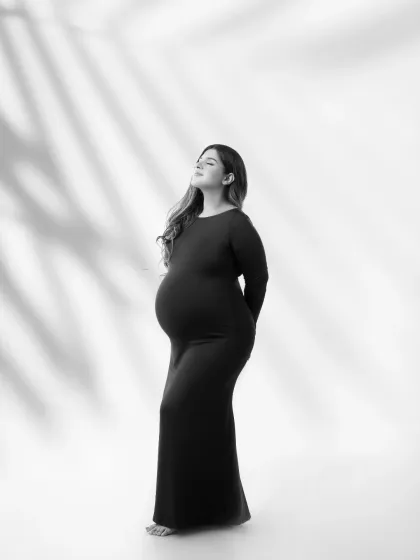 A serene and hopeful pose. This monochrome portrait uses soft, leaf-patterned shadows to add texture and artistry, as the mother-to-be looks up with a peaceful expression.