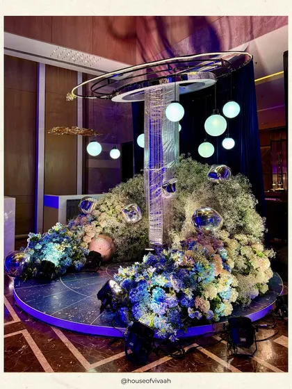 A sculptural centerpiece for the galaxy-themed Sangeet, combining a crystal fringe column with suspended orbs and a base of blue and white florals, creating a futuristic art piece.