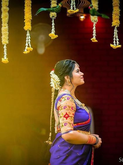 A bride in a blue saree, looking up at hanging floral decorations with a soft, happy expression.