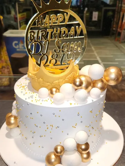 A cake fit for a king of the turntables. This elegant white and gold cake is decorated with a large crown and a custom "Happy Birthday DJ Sam" topper, perfect for a stylish celebration.