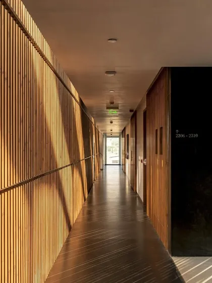 Light streams down a corridor with a slatted wooden wall, creating a rhythmic pattern of light and shadow that transforms a simple passageway into an experience.