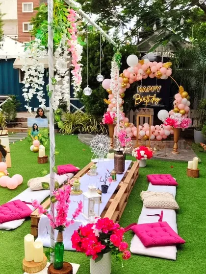 A beautiful garden picnic party in the daytime. The long pallet table is decorated with bright pink cushions and floral arrangements, with a stylish birthday backdrop and personalized photos completing the scene.