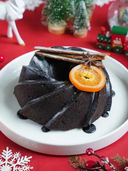 A dark chocolate bundt cake with a candied orange slice, set against a festive red background with snowflakes. The rich, moody styling makes this a sophisticated holiday dessert option.